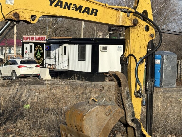Bear's Den Cannabis dispensary trailer with "No Tax" sign, viewed through a Yanmar excavator arm at a construction site.